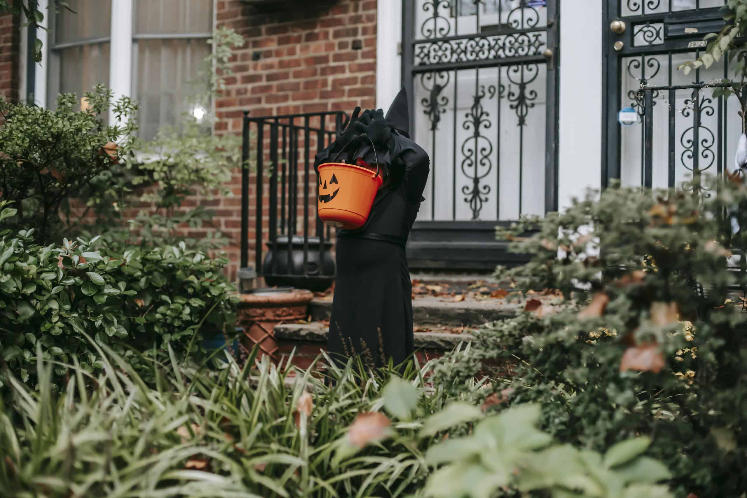 A child in a dark costume holding a pumpkin bucket stands at a house entrance during Halloween.