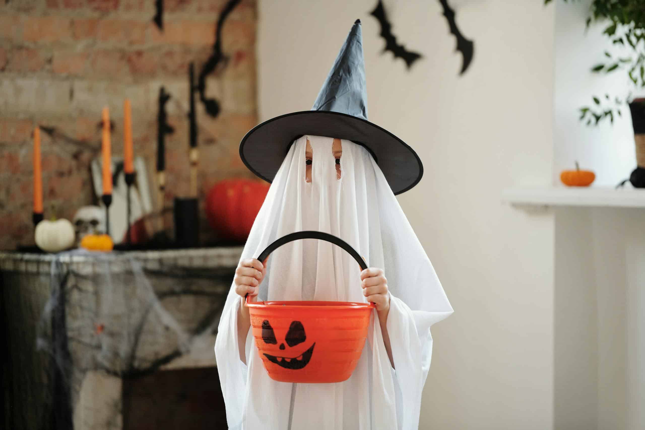 A child in a ghost Halloween costume holding a pumpkin basket, decorated room with spooky theme.