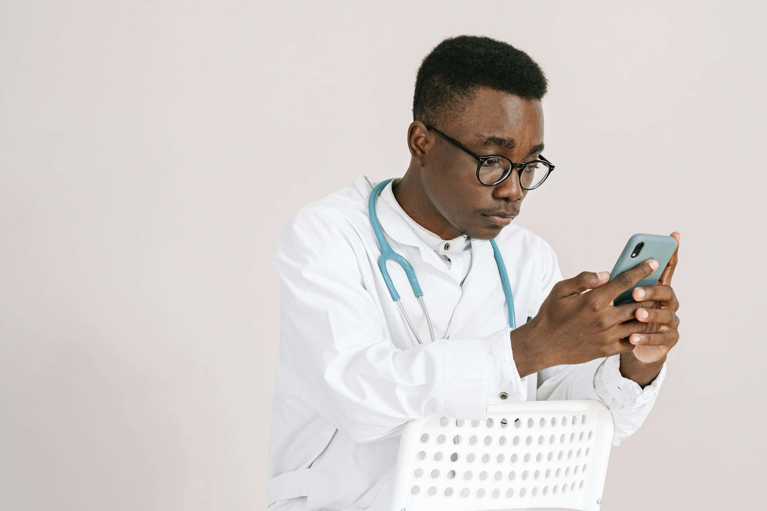 Young male doctor with stethoscope browsing on smartphone indoors.